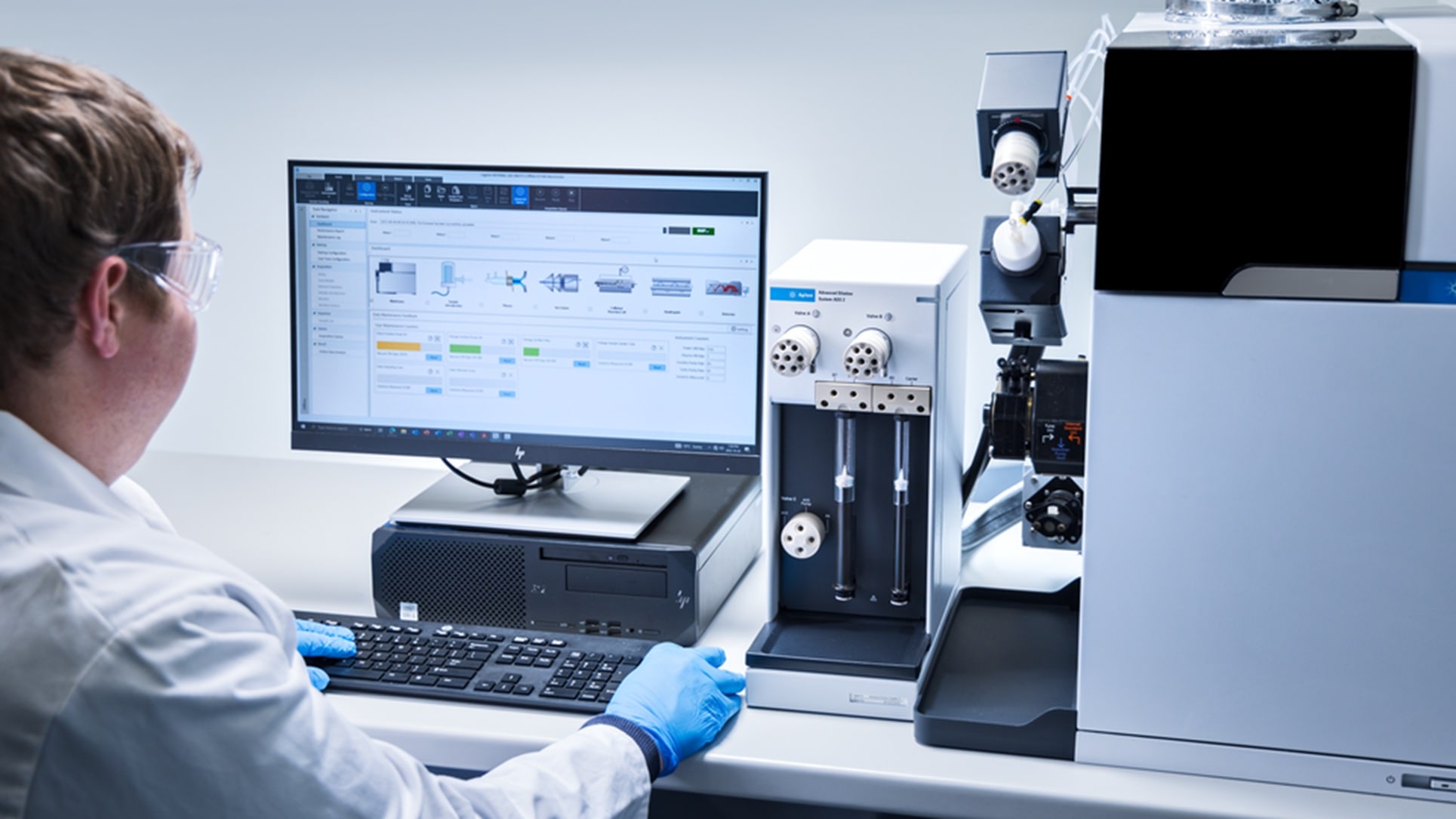 A scientist wearing safety goggles and blue gloves operates a computer connected to analytical lab equipment. The lab is orderly and high-tech.