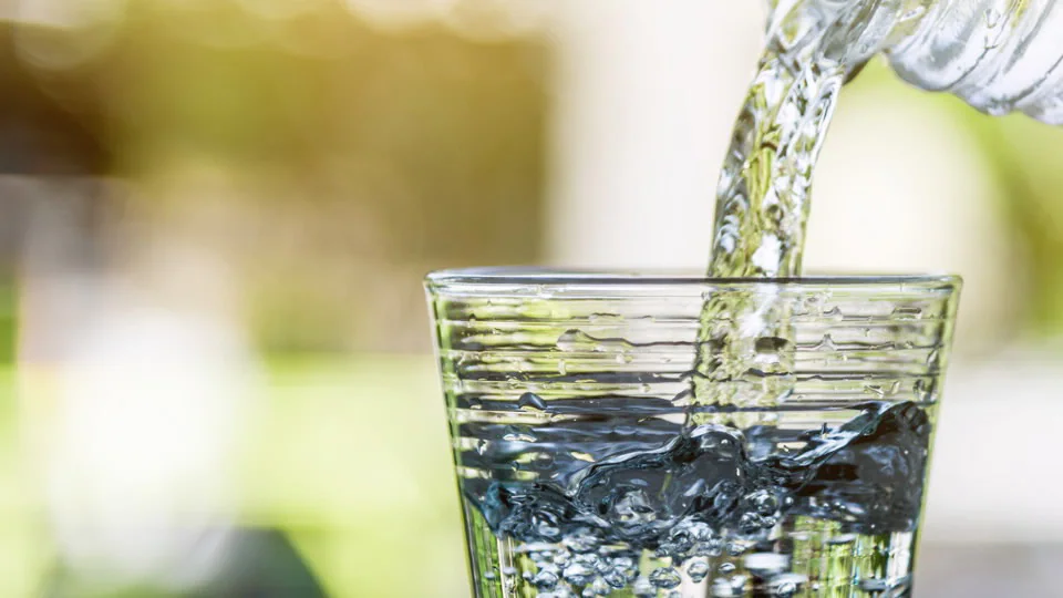 A clear glass is being filled with water from a plastic bottle, capturing splashes and bubbles, set against a softly blurred green and yellow background.